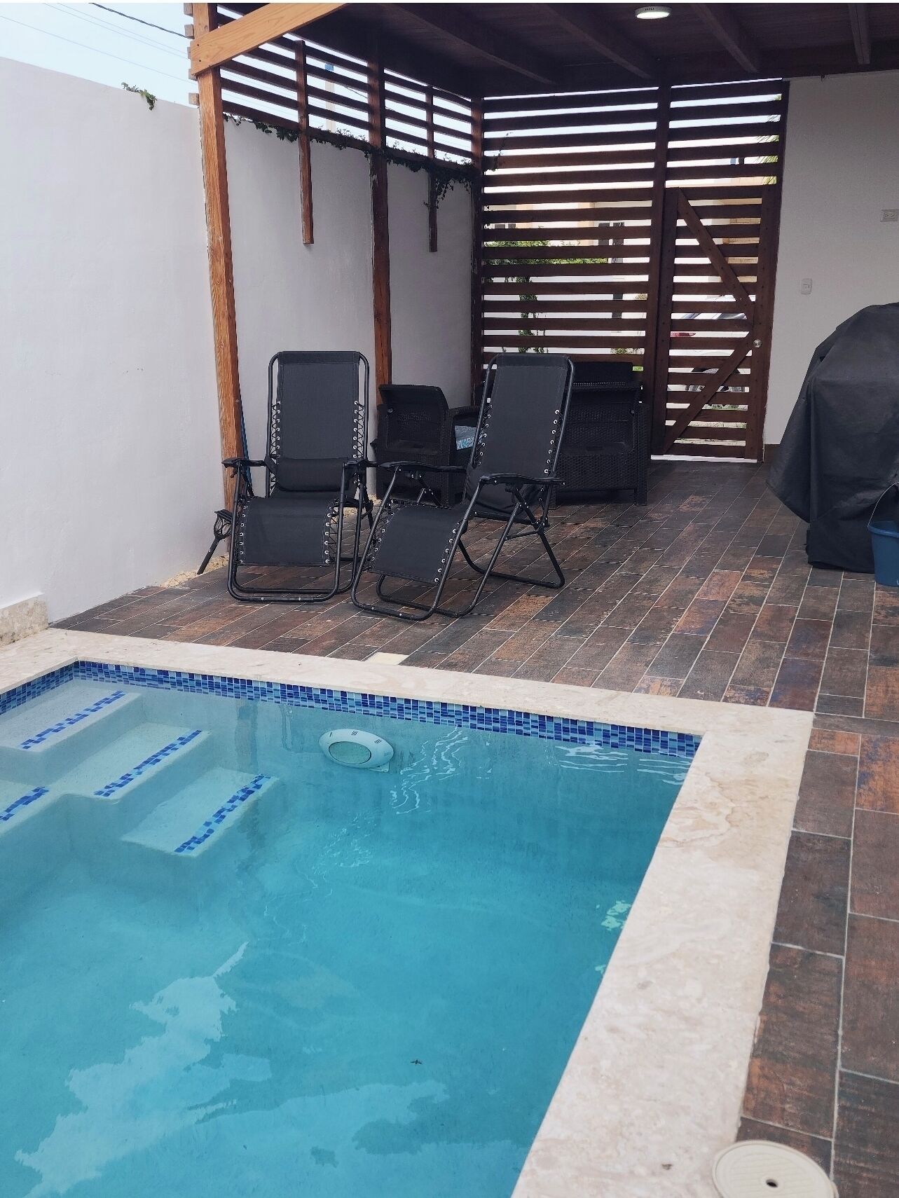 Una piscina con bordo piastrellato, circondata da una pergola in legno e poltroncine da relax nere.
