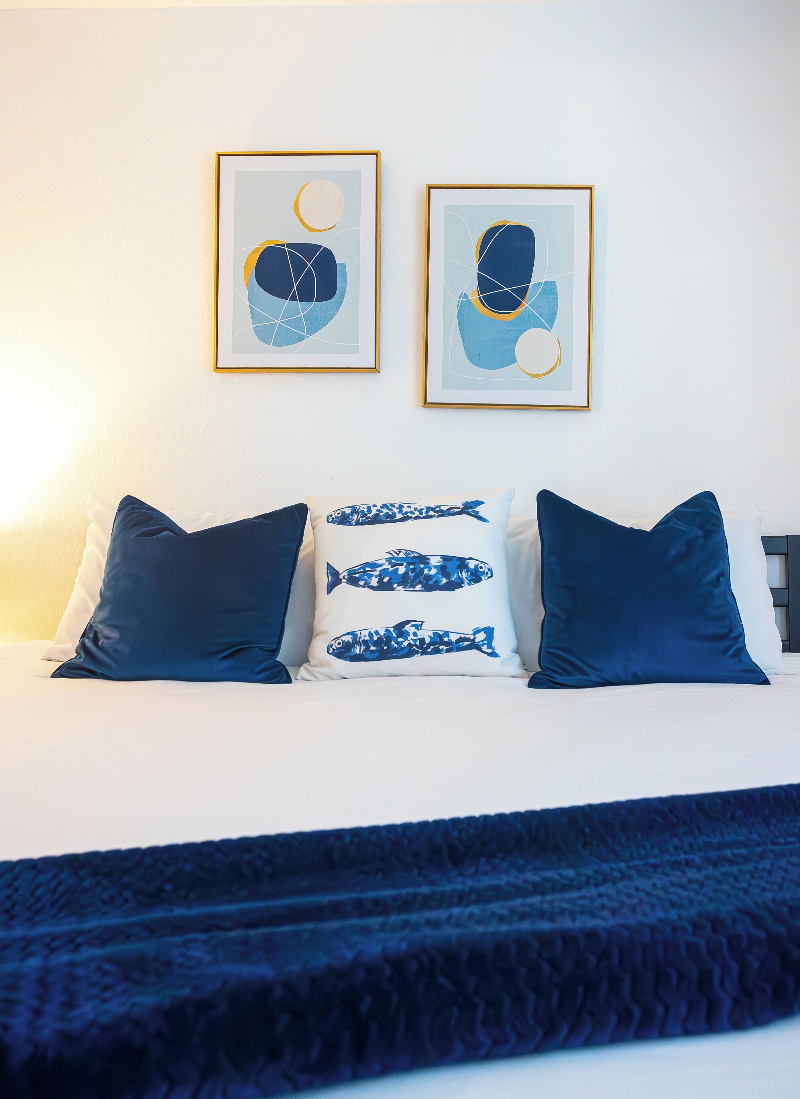 A bed with blue and white pillows, two framed abstract paintings on the wall, and a dark blue blanket.