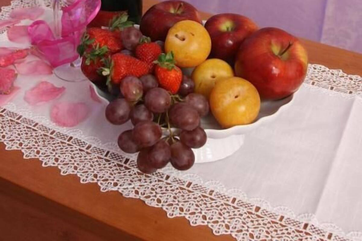 Une corbeille de fruits et une bouteille de champagne sont disposées avec soin sur une table recouverte d’une nappe en dentelle. À côté, on aperçoit une table de chevet en bois avec une lampe, et un lit recouvert d’une courtepointe blanche, le tout dans une pièce aux murs violets.