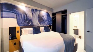 Una habitación de hotel moderna con una cama grande, una obra de arte colgada en la pared, una mesita de noche con una botella de agua y un armario.