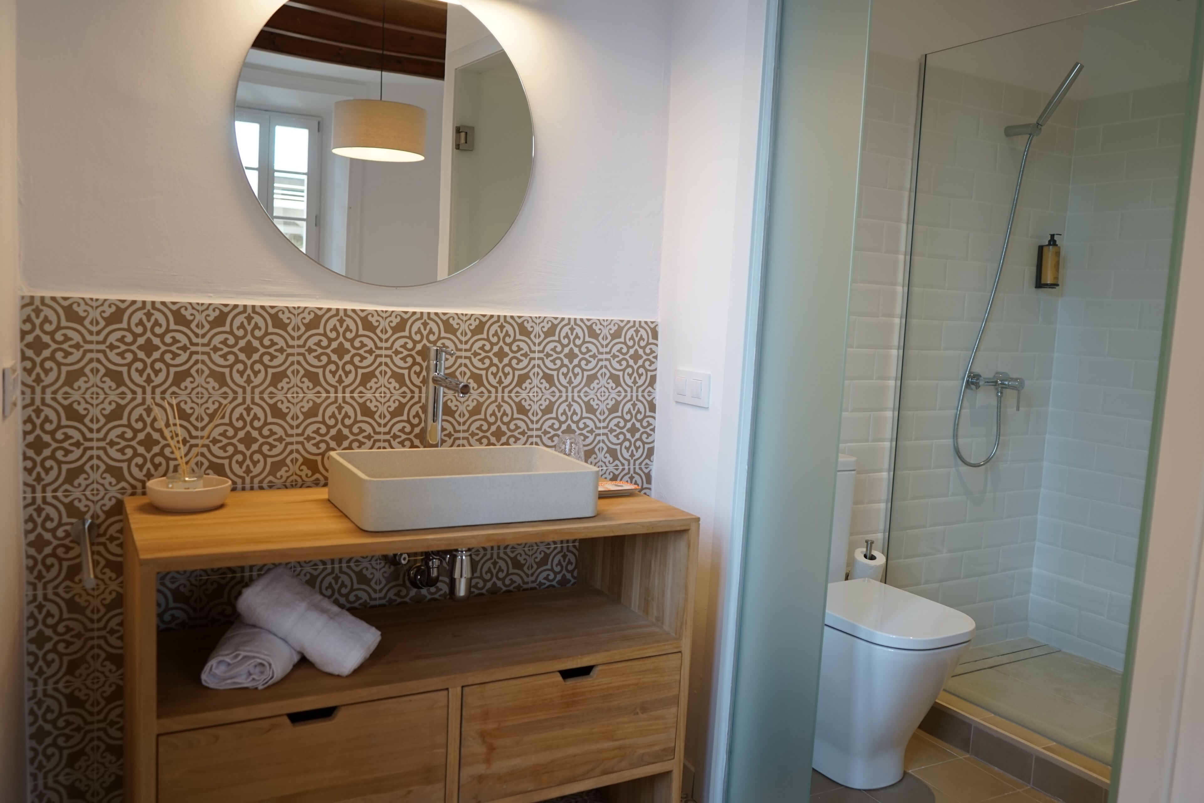 A bathroom with a wooden vanity, a round mirror, patterned tiles, a shower enclosure, and a toilet.