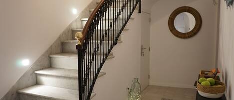 A staircase with marble steps and a black metal railing, a round mirror on the wall, a patterned rug, and a basket with green apples on a small table.