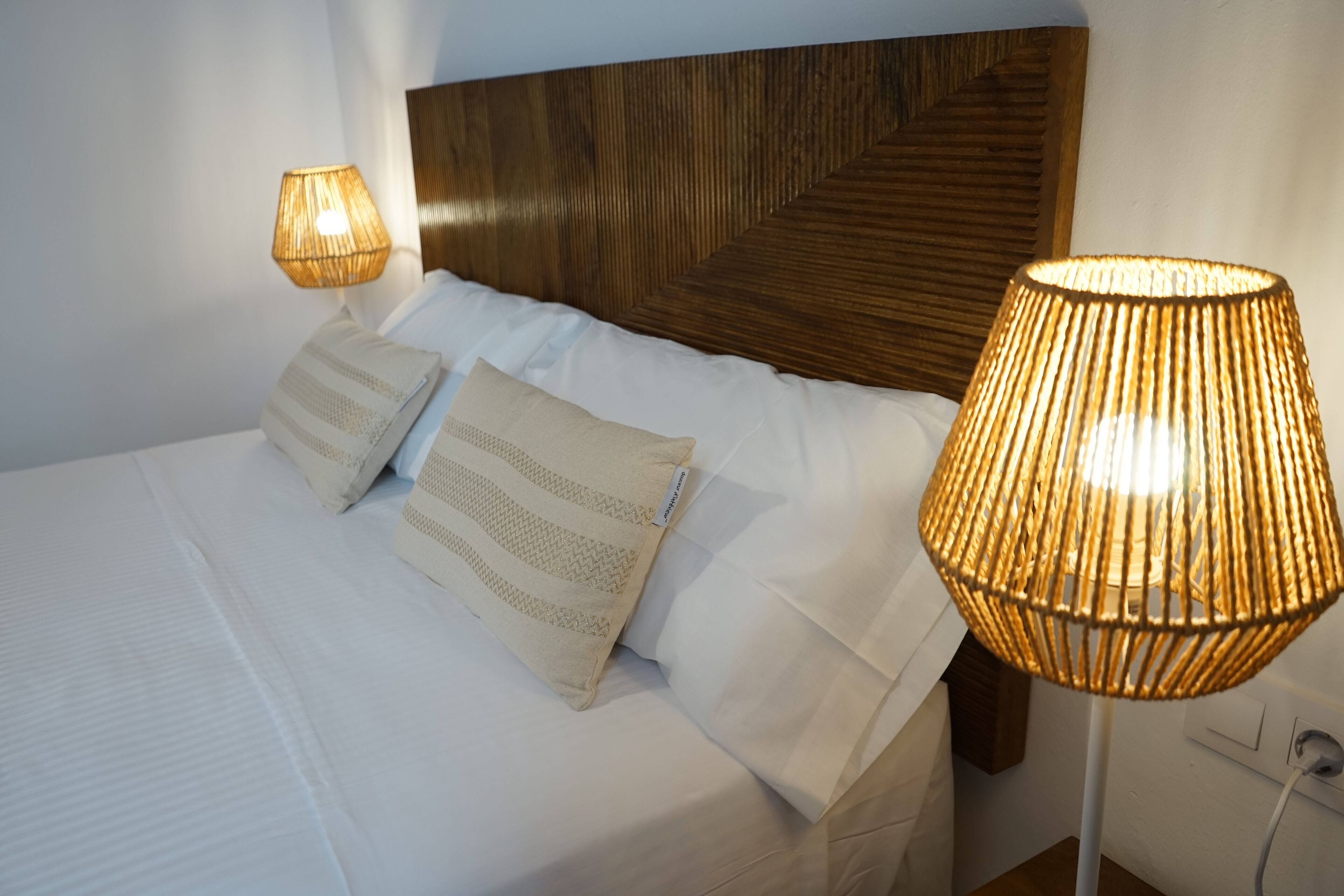 A neatly made bed with white linens and two decorative pillows, a wooden headboard, and two wall-mounted lamps with a woven design.