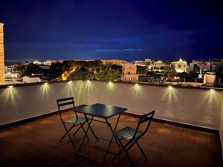 A rooftop terrace with a table and chairs, overlooking a cityscape at night.