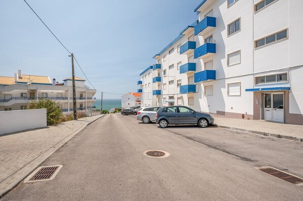 A residential street with apartment buildings, a parked car, and a clear view of the sea. - GuestReady - Floral Ericeira residence (Mafra)