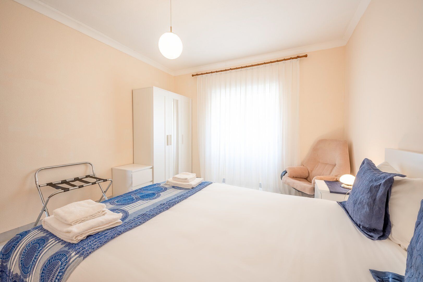 A hotel room with a bed, a chair, a wardrobe, and a window with curtains.