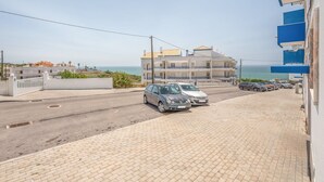 Un estacionamiento con dos coches aparcados frente a un edificio de fachada azul y blanca, con vista a un cuerpo de agua.