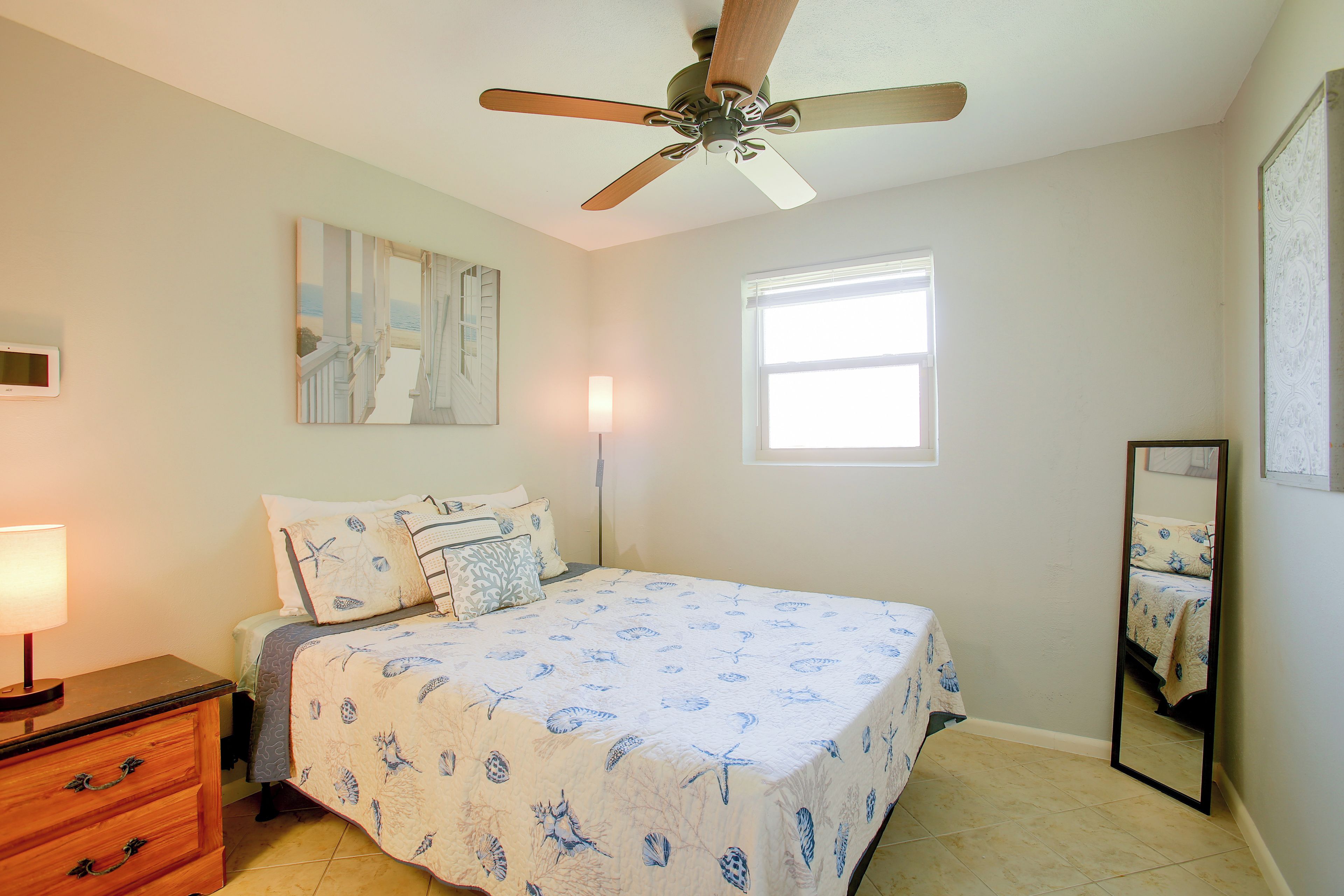 A bedroom with a bed, a nightstand, a ceiling fan, a window, and a wall-mounted mirror.