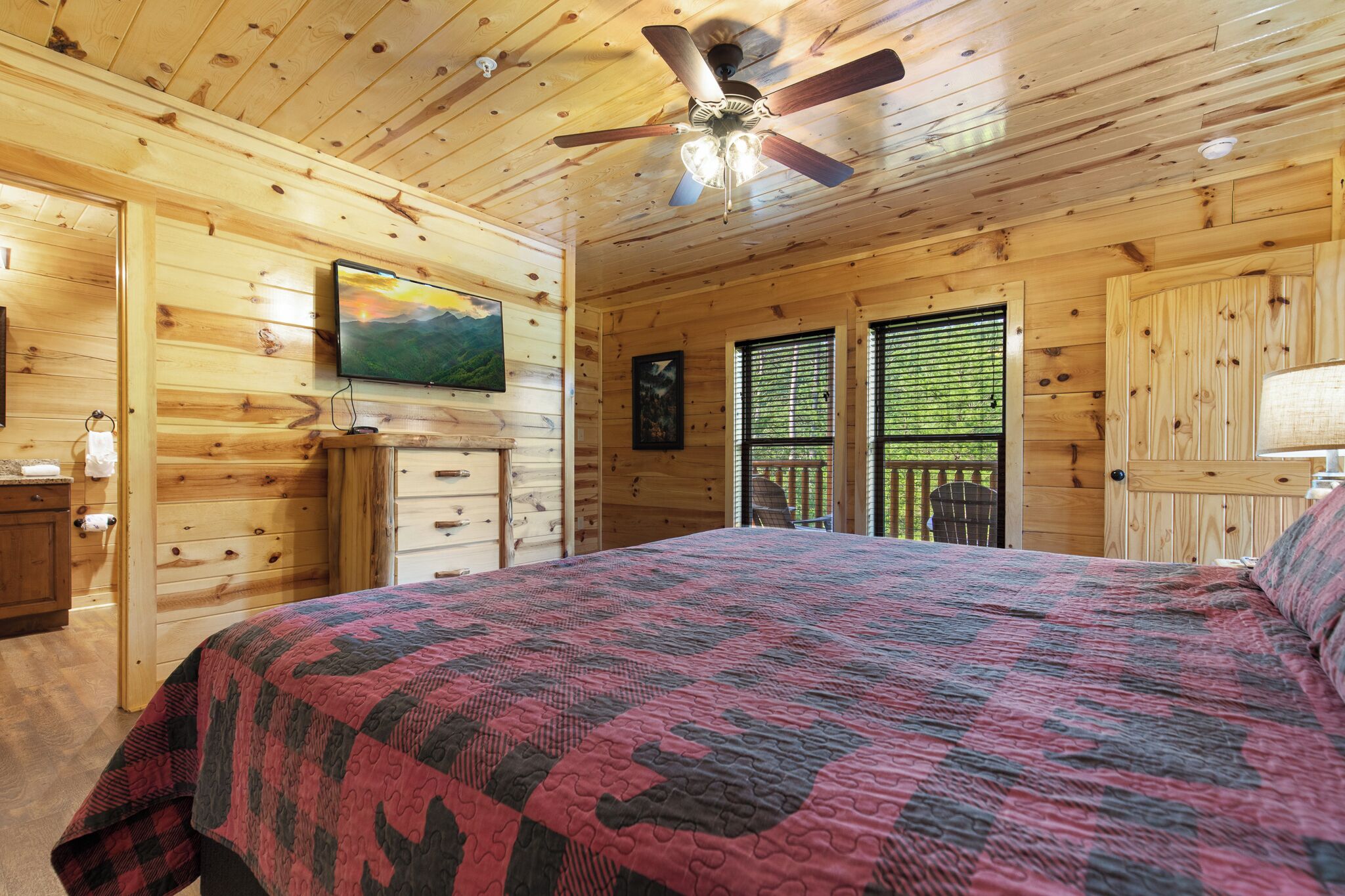 A wooden cabin bedroom with a bed, a ceiling fan, a TV, and a window with blinds.