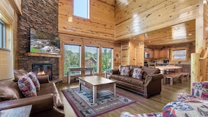A spacious log cabin interior with a stone fireplace, wooden furniture, and large windows. - Mountain High Cinema by Jackson Mountain Rentals (Sevierville)