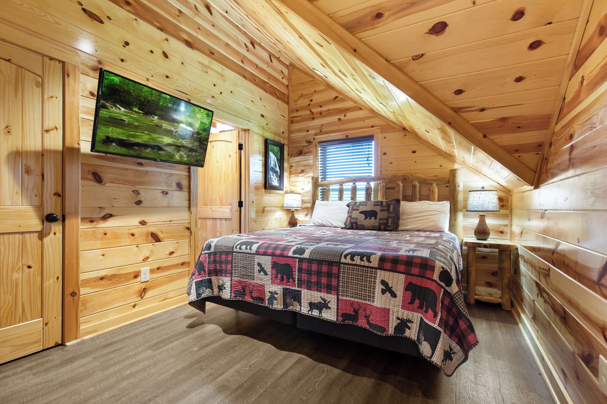 A cozy cabin room with a wooden interior, a bed with a quilt, a television mounted on the wall, and a window with blinds.