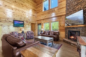A log cabin interior with a fireplace, wooden walls, and a large window overlooking a scenic landscape. - Mountain High Cinema by Jackson Mountain Rentals (Sevierville)