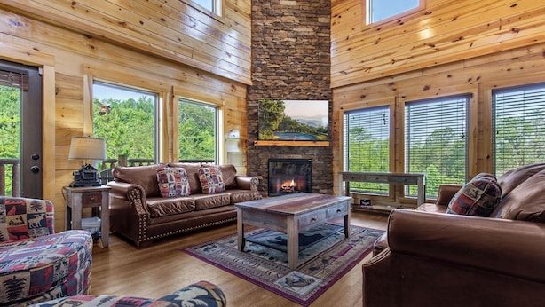 A cozy living room with wooden walls, a stone fireplace, and large windows offering a view of the outdoors. - Mountain High Cinema by Jackson Mountain Rentals (Sevierville)