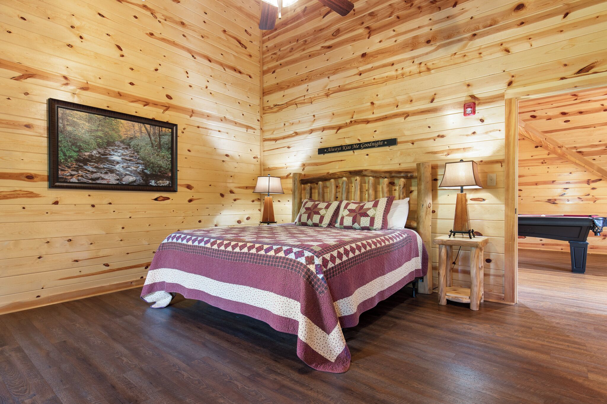 A wooden cabin bedroom with a bed, two bedside lamps, a framed picture on the wall, and a sign above the bed.