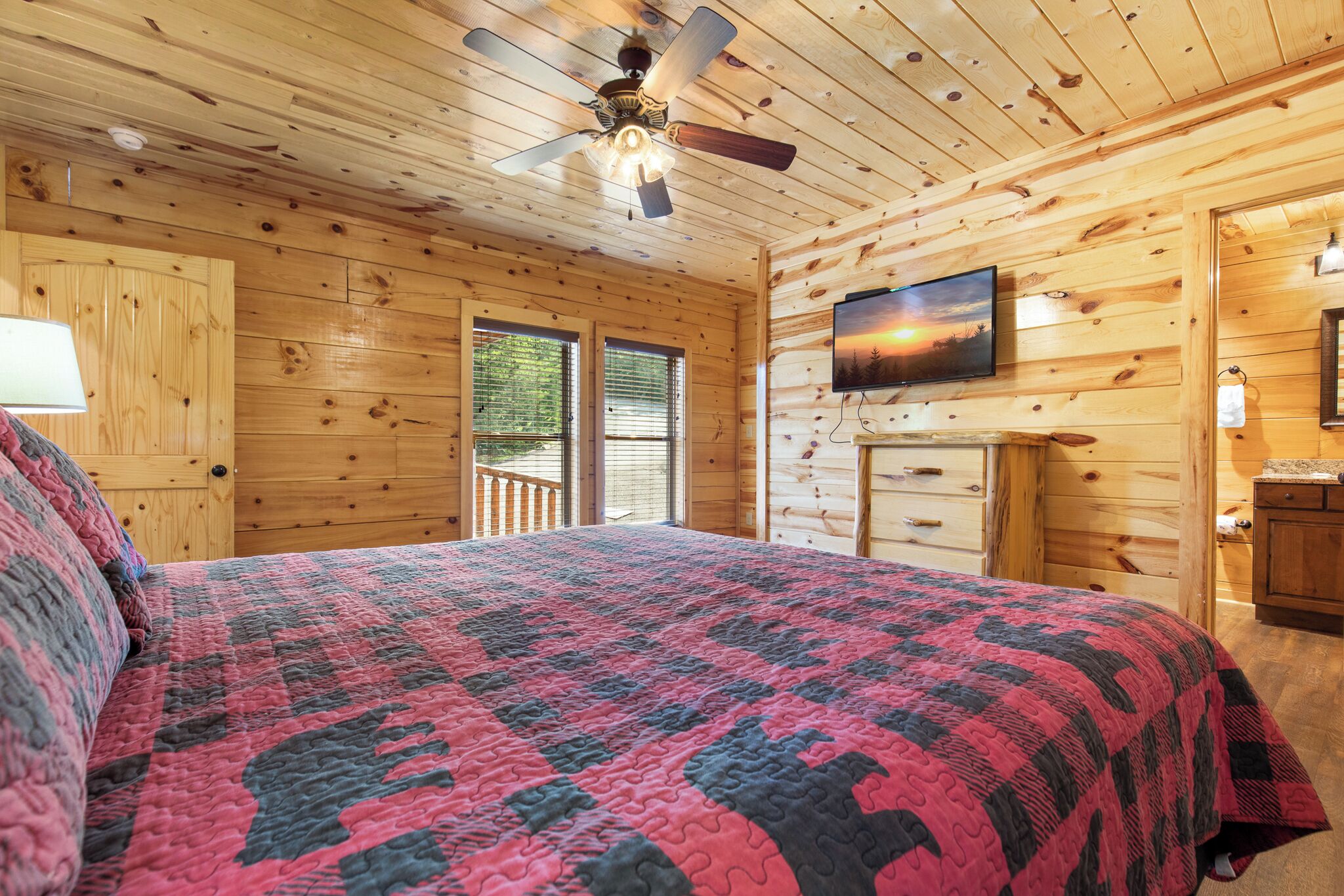 Quarto de cabana de madeira com uma cama, um ventilador de teto, uma TV fixada na parede e uma janela.