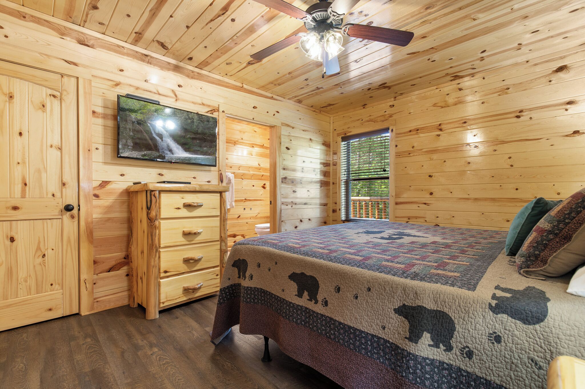 A wooden cabin bedroom with a bed, a TV mounted on the wall, a ceiling fan, and a window with blinds.