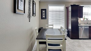 A dining area with a table and chairs, a kitchen with cabinets and appliances, and a window with blinds. - Beach Daze by Ryson Vacation Rentals (Galveston)