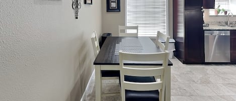 Un coin repas avec une table et des chaises, une cuisine équipée de placards et d’appareils électroménagers, et une fenêtre avec des stores.