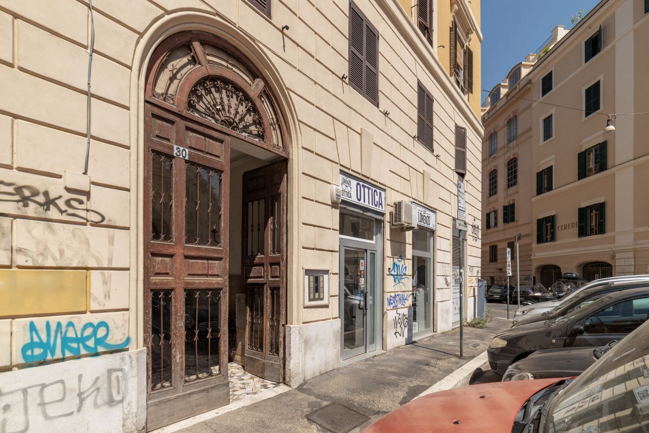 A building with a large arched entrance, a shop named 'Ottica,' and a street with parked cars.