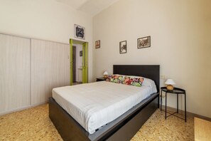 A bedroom with a bed, a nightstand, a lamp, a door, and a wall with framed pictures.