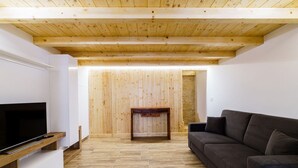 Ein modernes Wohnzimmer mit Holzbalkendecke, einem Flachbildfernseher, einem dunklen Sofa und einem kleinen Holztisch.