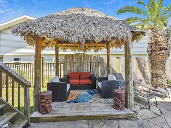 A covered outdoor seating area with a thatched roof, wicker furniture, and a decorative rug. - Tiki Lagoon by Ryson Vacation Rentals (Galveston)