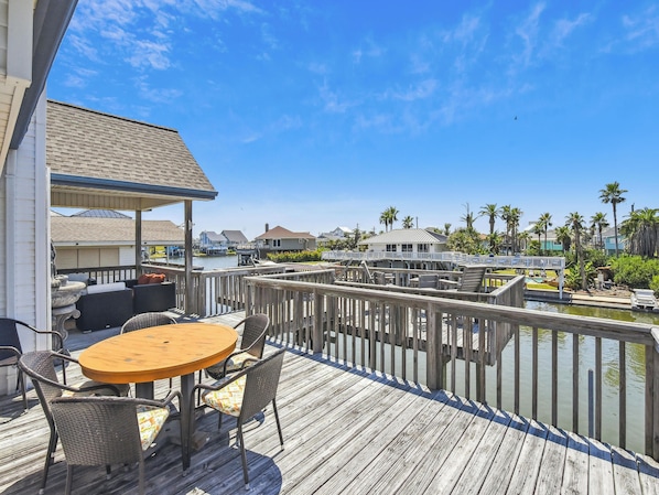 A wooden deck with a round table and chairs, overlooking a marina with boats and houses in the background.