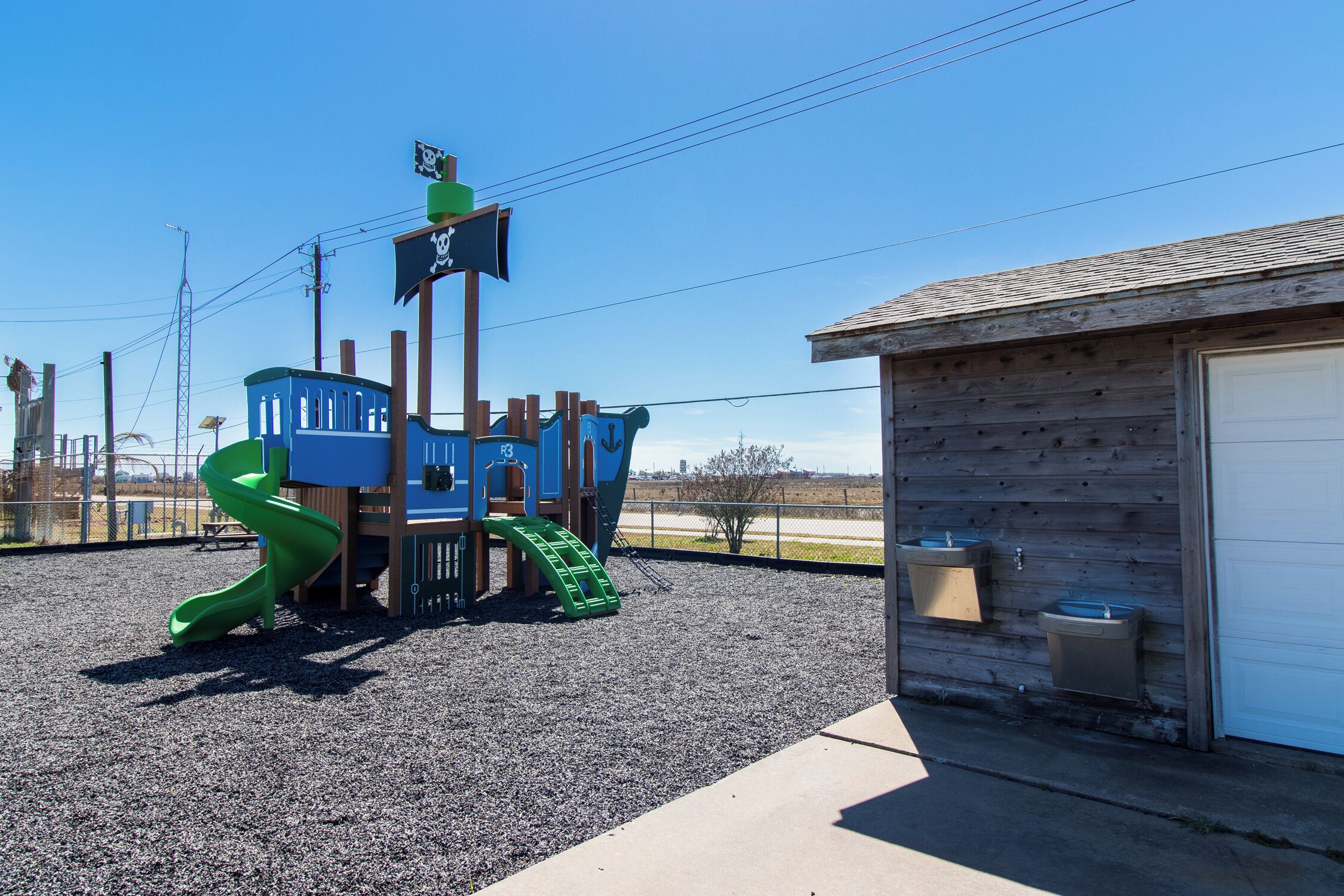 A playground with a slide and climbing structures, a small building with a garage door, and a gravel ground.