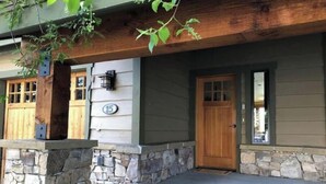 A two-story house with a stone foundation, wooden siding, and a front porch with steps leading to a wooden door. - Cozy Home steps from Town Center, with a Stunning View of the Mountains! (Big Sky)