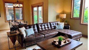 A spacious living room with a large brown leather sectional sofa, a wooden coffee table, and a dining area with a table and chairs. - Cozy Home steps from Town Center, with a Stunning View of the Mountains! (Big Sky)