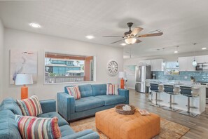 A modern living room with a blue sofa, an orange ottoman, and a dining area with bar stools. - After Dune Delight by Silver Sands Vacation Rentals (Port Aransas)