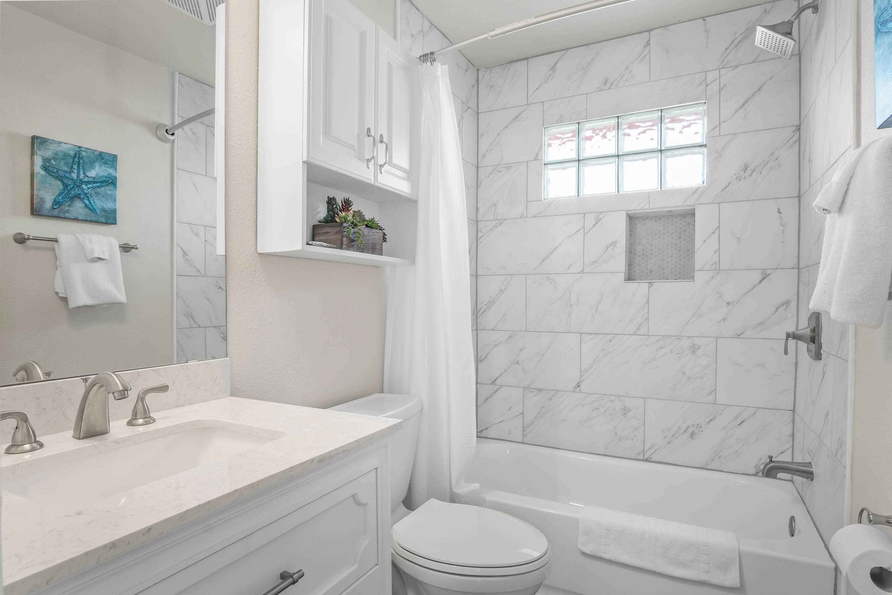 A bathroom with a white sink, toilet, and bathtub, marble walls, and a window with a view.