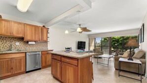 A spacious kitchen with wooden cabinets, a built-in dishwasher, and a large island. There is a dining area with a sofa and a coffee table. - Kihei Akahi D-104 (Kihei)
