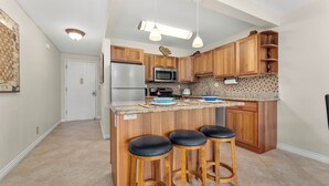 A kitchen with wooden cabinets, a stainless steel refrigerator, a microwave, and a granite countertop. There are three stools with black seats. - Kihei Akahi D-104 (Kihei)