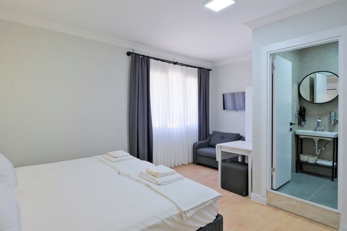 Une chambre d’hôtel moderne dotée d’un grand lit, d’un bureau, d’une chaise, d’une salle de bain avec un lavabo et un miroir, et d’un téléviseur fixé au mur.