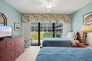 A hotel room with a bed, a TV, a dresser, and a balcony with a view of a building and a grassy area.