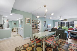 A modern kitchen with white cabinets, a dining area with a glass table and colorful chairs, and a living room with a sofa and a TV.