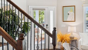 A staircase with a wooden handrail and black balusters, a white wall with a framed artwork, a white chair, a table with a lamp, and a door leading outside. - 2-bedroom vacation home just a short walk to Rosemary Beach (Rosemary Beach)