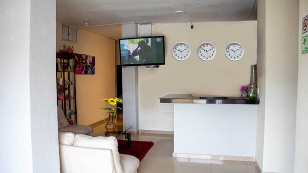 A living room with a sofa, a TV, three wall clocks, and a kitchen area with a counter and sink. - Hotel Ruta Del Mar (Santa Marta)