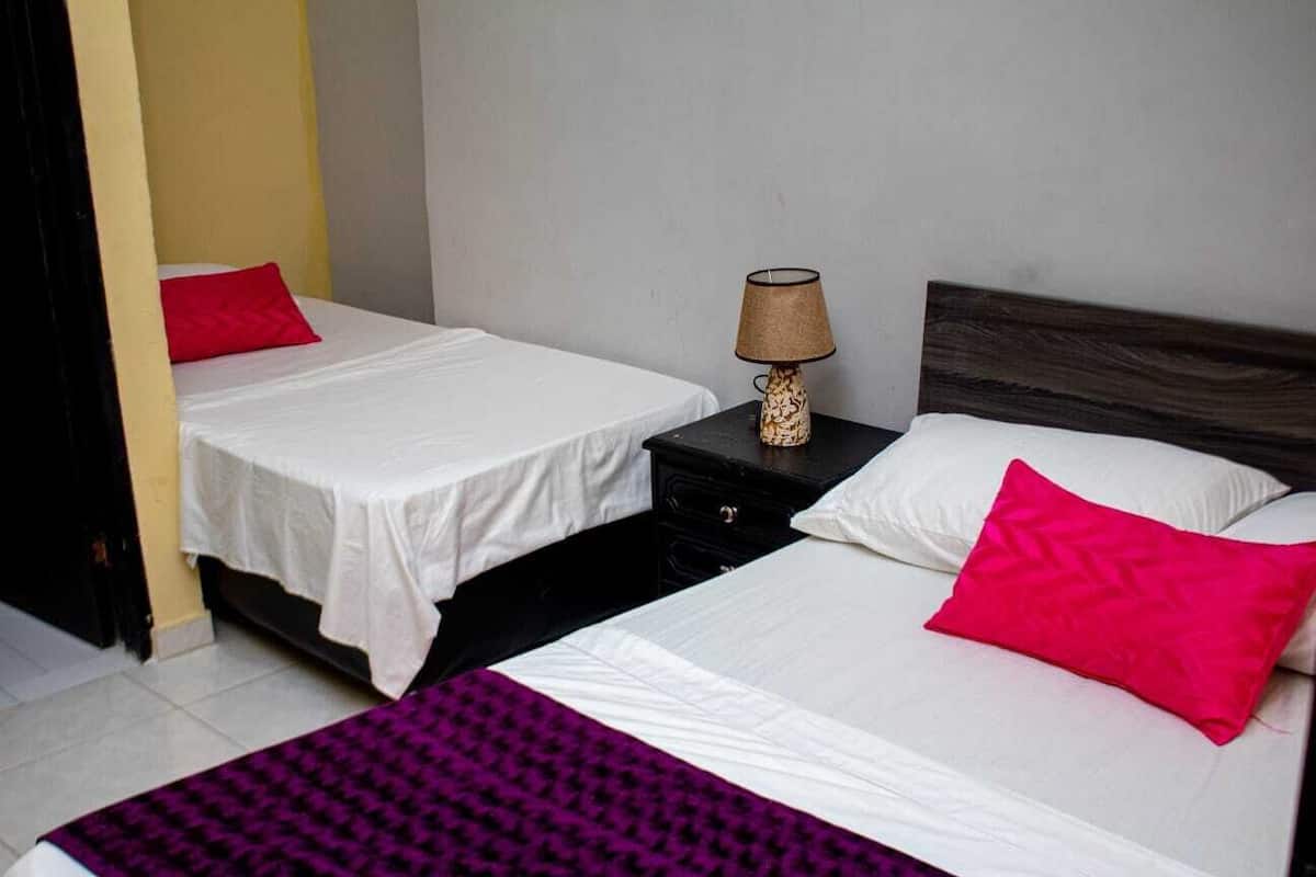 A hotel room with two beds, each with white bedding and a red pillow. There is a nightstand with a lamp between the beds.