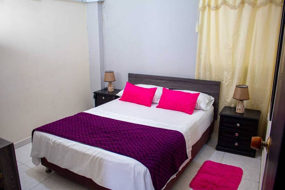 A bed with a white and purple bedspread, two pink pillows, and two nightstands with lamps.
