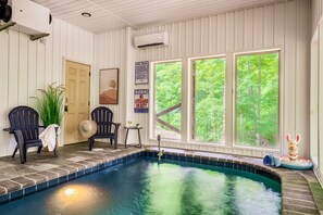 Une piscine intérieure avec vue sur les arbres, deux fauteuils Adirondack, un ventilateur et un panneau indiquant la présence d’un sauveteur.