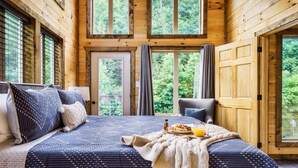 Ein Schlafzimmer einer Holzhütte mit einem großen Bett, einem Frühstücksbrett und Blick auf den Wald durch die Fenster.