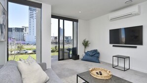 Ein modernes Wohnzimmer mit grauem Sofa, einem kleinen Couchtisch, einem an der Wand montierten Fernseher und Blick auf eine Stadtlandschaft durch große Fenster.