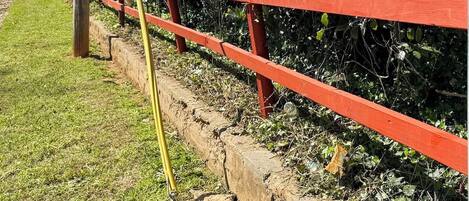 A red metal fence with a yellow pole, a concrete wall, and a grassy area with bushes.