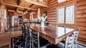 A log cabin interior with a large wooden dining table, chairs, a chandelier, and a kitchen area with appliances. - 7BR LogCabin/Spa/Nature/MontTremblant Area/18 Guests  (Labelle)
