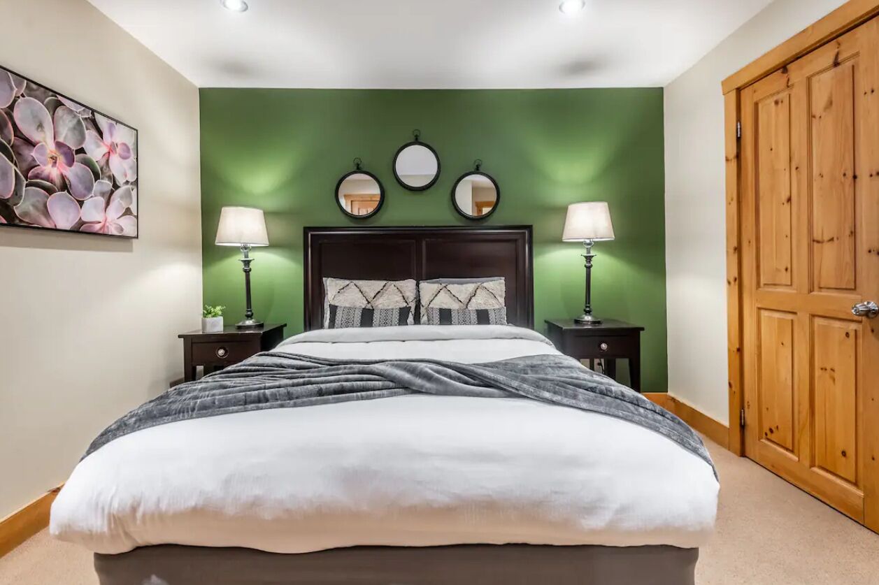 Quarto com cama, duas mesas de cabeceira com abajures, uma parede verde e uma porta de madeira.