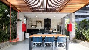 An outdoor dining area with a wooden table and blue chairs, a black grill, and red cabinets. - New studio in Pinheiros - Great Location - comfort - safety (Pinheiros São Paulo)