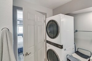 Une buanderie équipée d’une machine à laver et d’un sèche-linge superposés, d’un étendoir métallique et d’une porte donnant sur une chambre à coucher avec un lit et une fenêtre.