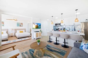 Quarto de hotel moderno com cama, mesas de cabeceira, televisão, área de cozinha com banquetas e mesa de jantar.